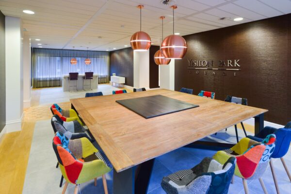 Moderne lounge in Ypsilon Park met grote houten vergadertafel, kleurrijke stoelen en koperkleurige hanglampen.